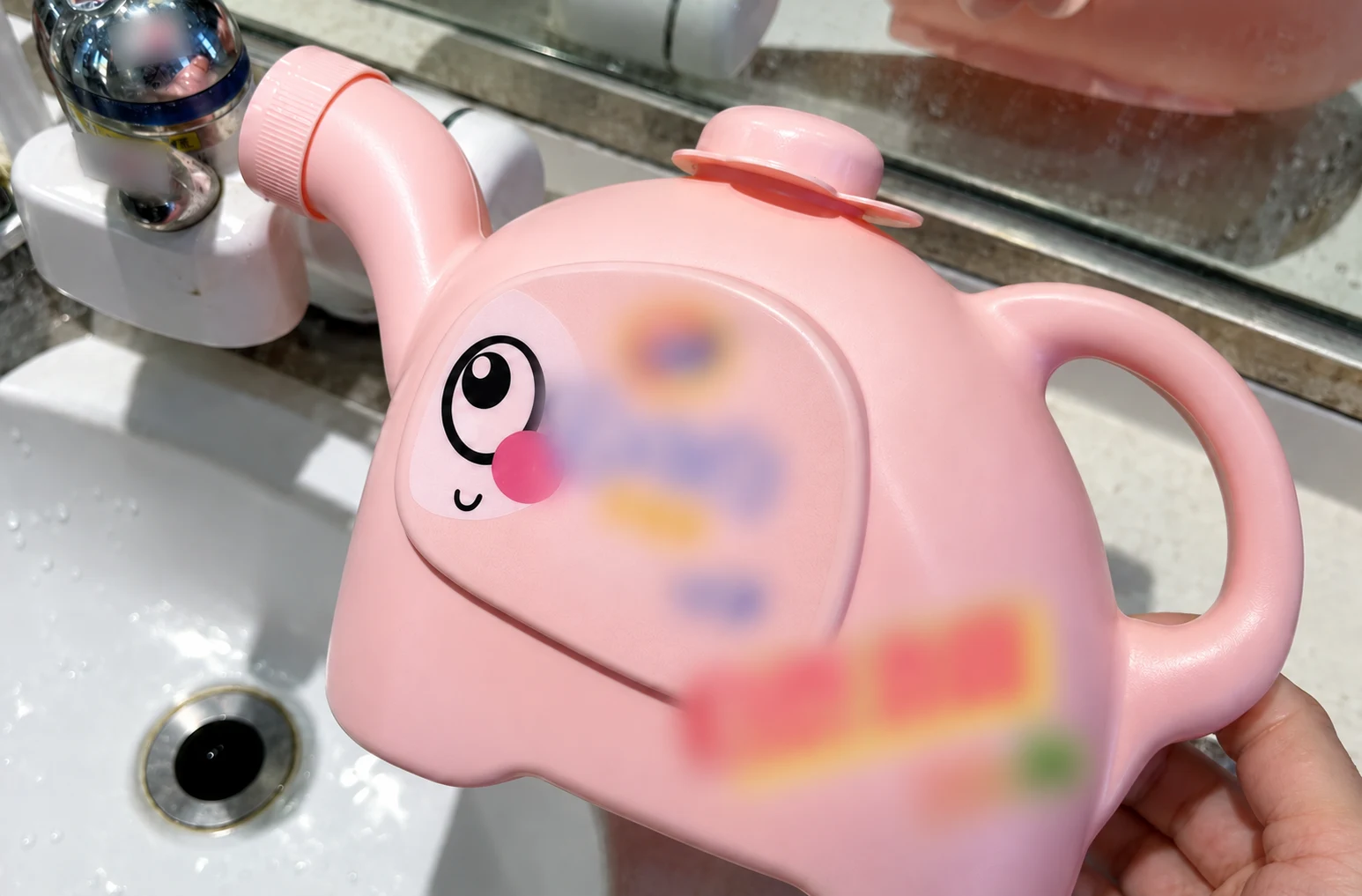 流し台のそばにあるピンク色のじょうろ型容器の近接画像。持ち手と注ぎ口が見え、商業的な文字は柔らかくぼかされています。